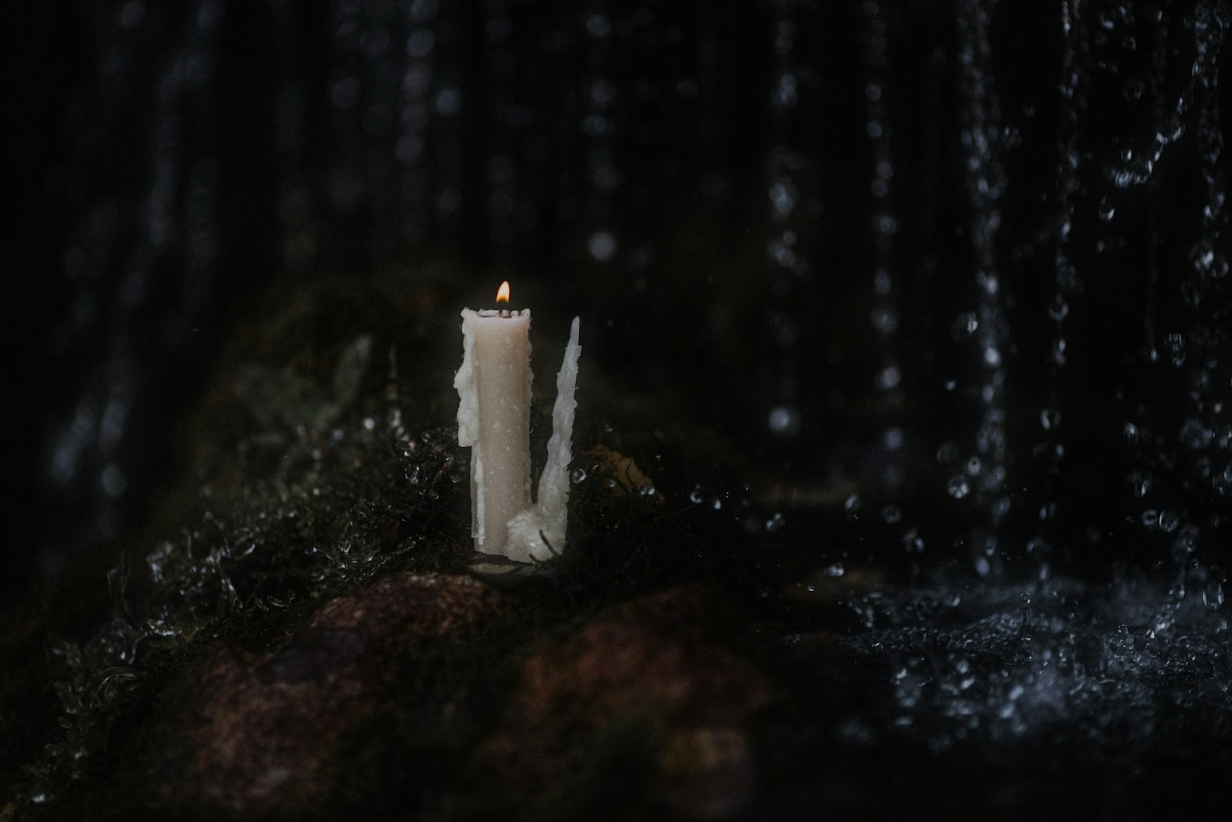 A single lit white candle sits on moss-covered rocks in the dark, with water droplets falling nearby and a faint stream flowing around it.